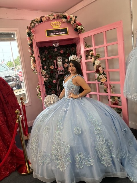 Quinceañera in a light blue ball gown posing at the pink flower-covered House of Quinceañeras photo door