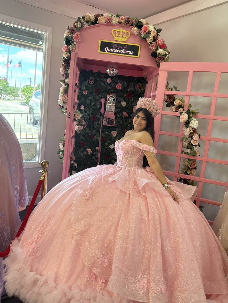 Quinceañera in a blush pink ball gown seated in front of the pink phone booth covered in roses