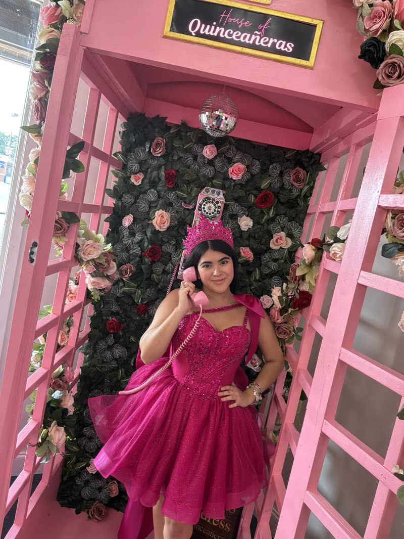 Quinceañera in a fuchsia dress posing inside the pink phone booth with a flower wall
