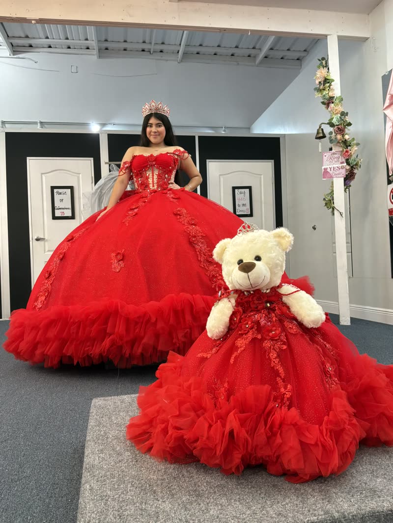 Quinceañera in a red ball gown and crown beside a teddy bear in a matching red dress