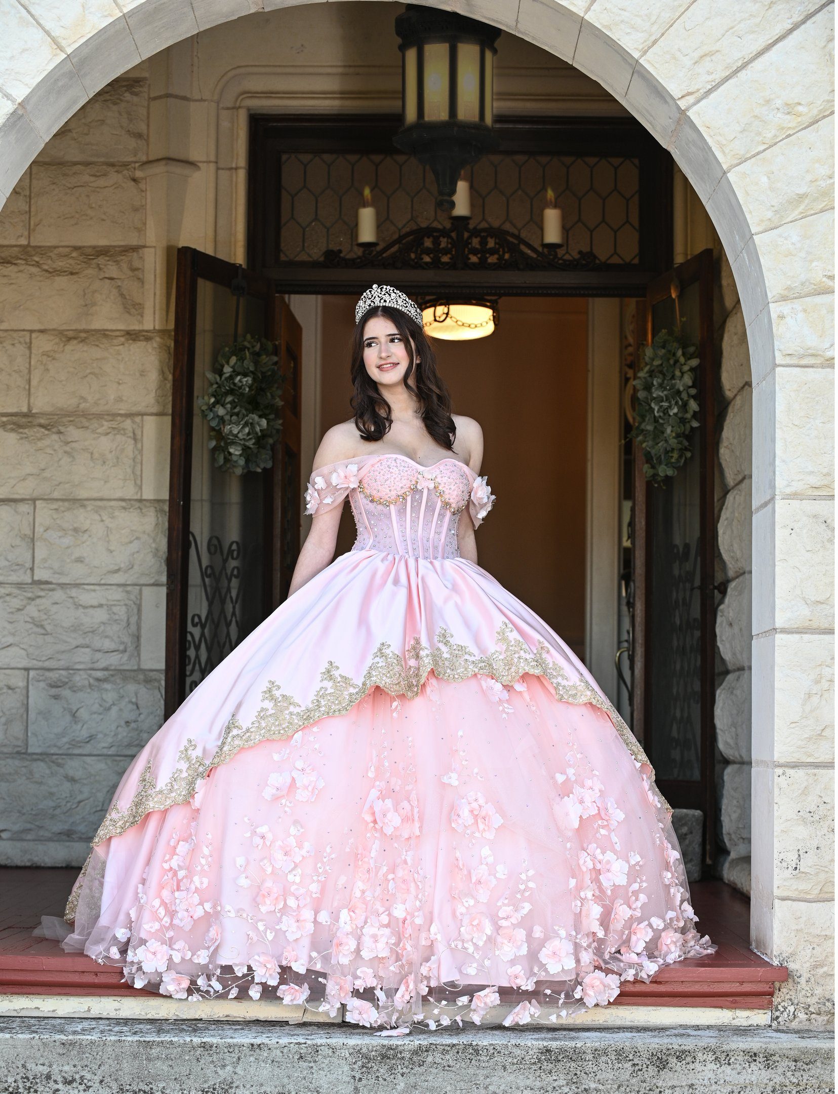 Quinceañera in a pink and gold ball gown with a tiara standing in a castle archway